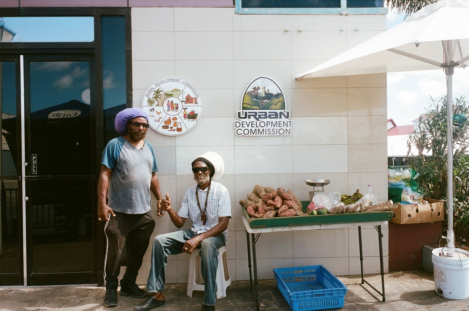 Two men at a roadside stall of sweet potatoes, yams and bananas outside offices carrying the signs of the Rural Development Commission and the Urban Development Commission